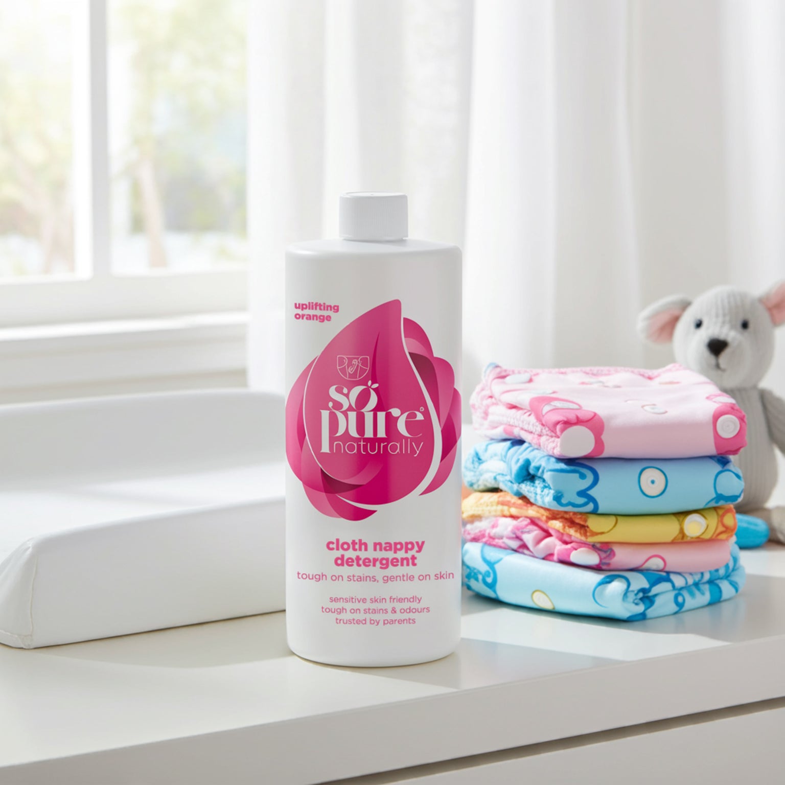 Bottle of 'so pure' cloth nappy detergent with folded colorful nappies on a white surface.